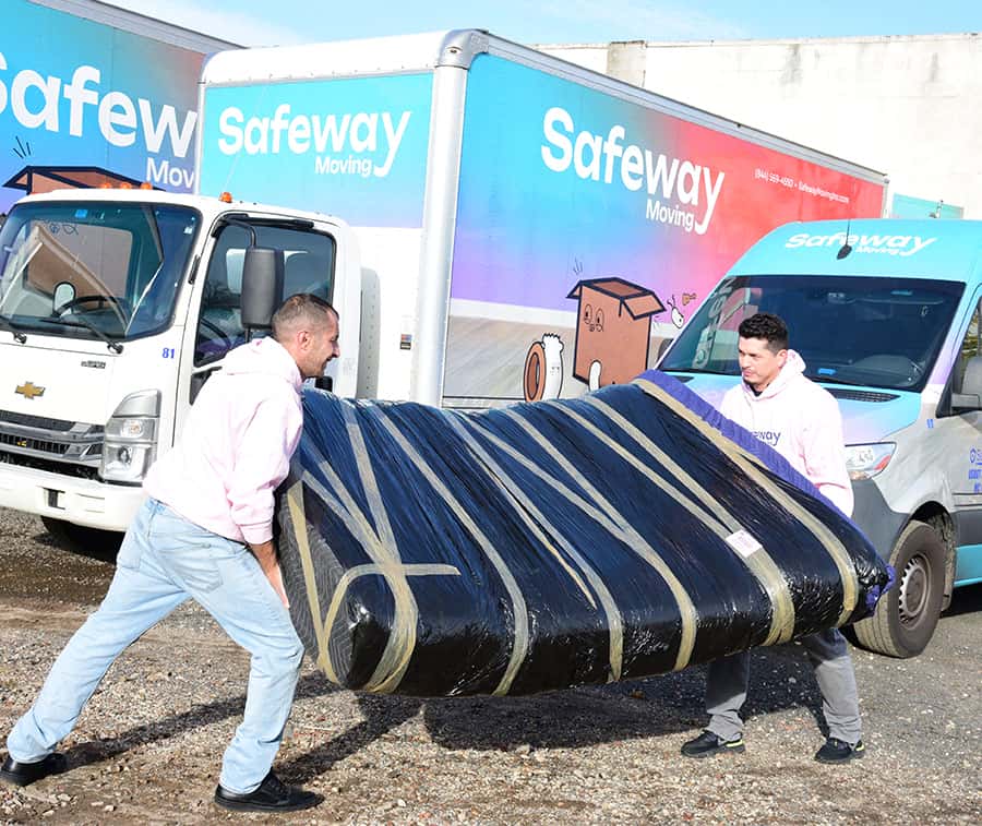 Two Safeway movers carrying a shrink-wrapped sofa past branded trucks